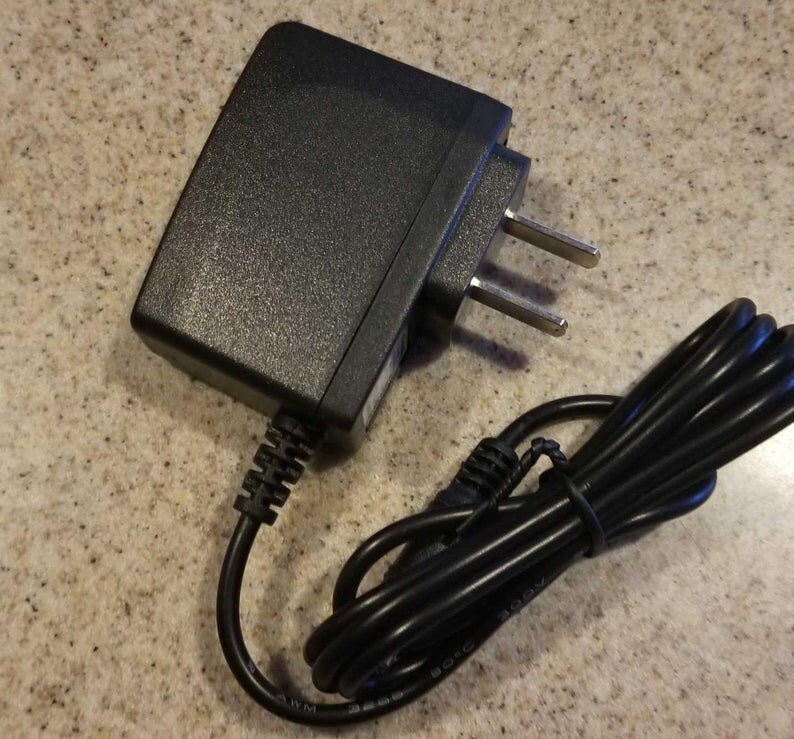 May include: A black AC power adapter with a two-prong plug and a black cord. The adapter is rectangular with a textured surface. The cord is coiled and secured.