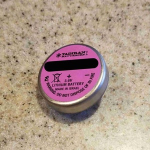 May include: A small, silver lithium battery with a pink top. The top has black text and the brand name "Tadiran Batteries". The battery has a warning label and the text "Lithium Battery Made in Israel".
