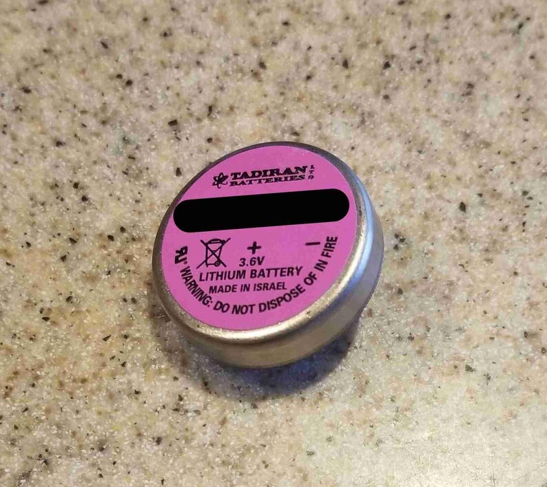 May include: A small, silver lithium battery with a pink top. The top has black text and the brand name "Tadiran Batteries". The battery has a warning label and the text "Lithium Battery Made in Israel".