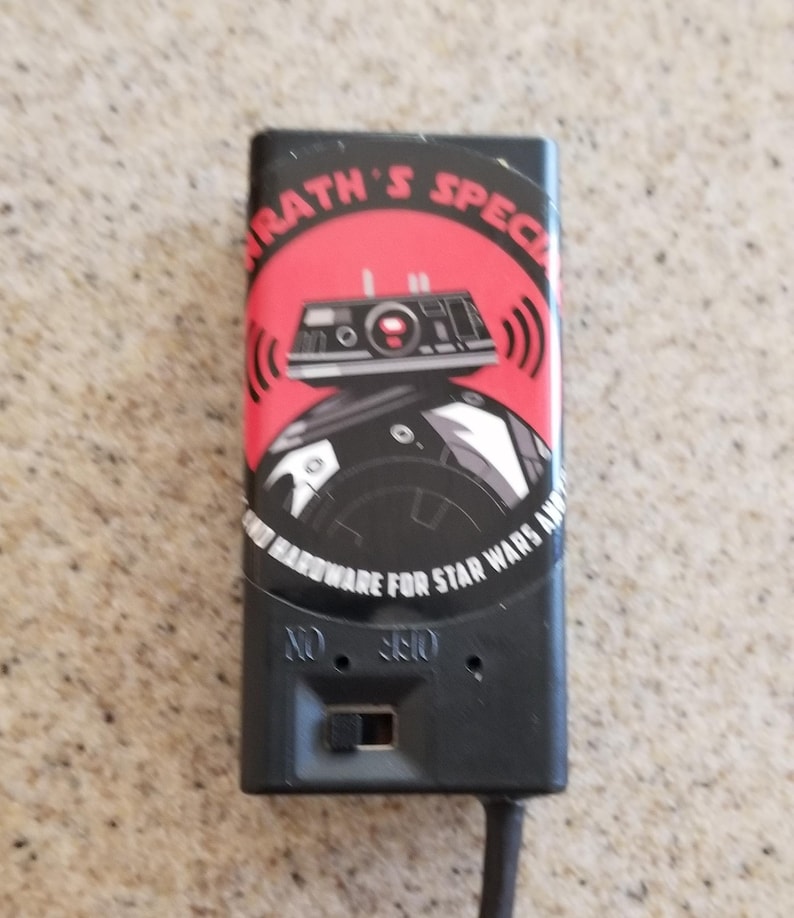 May include: Black rectangular device with a red and black graphic of a Star Wars droid and the text "KRATH'S SPECIAL HARDWARE FOR STAR WARS AMPS". The device has an on/off switch and a cord.