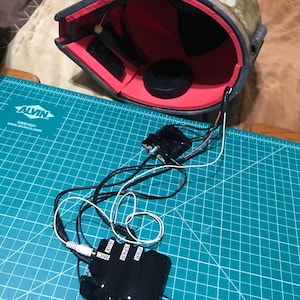 May include: A tan and red helmet interior with black wiring and electronic components. The helmet has a weathered appearance with red padding and a black interior. The wiring is connected to a black box with labels for MIC, AUX, SPKR, and PWR.