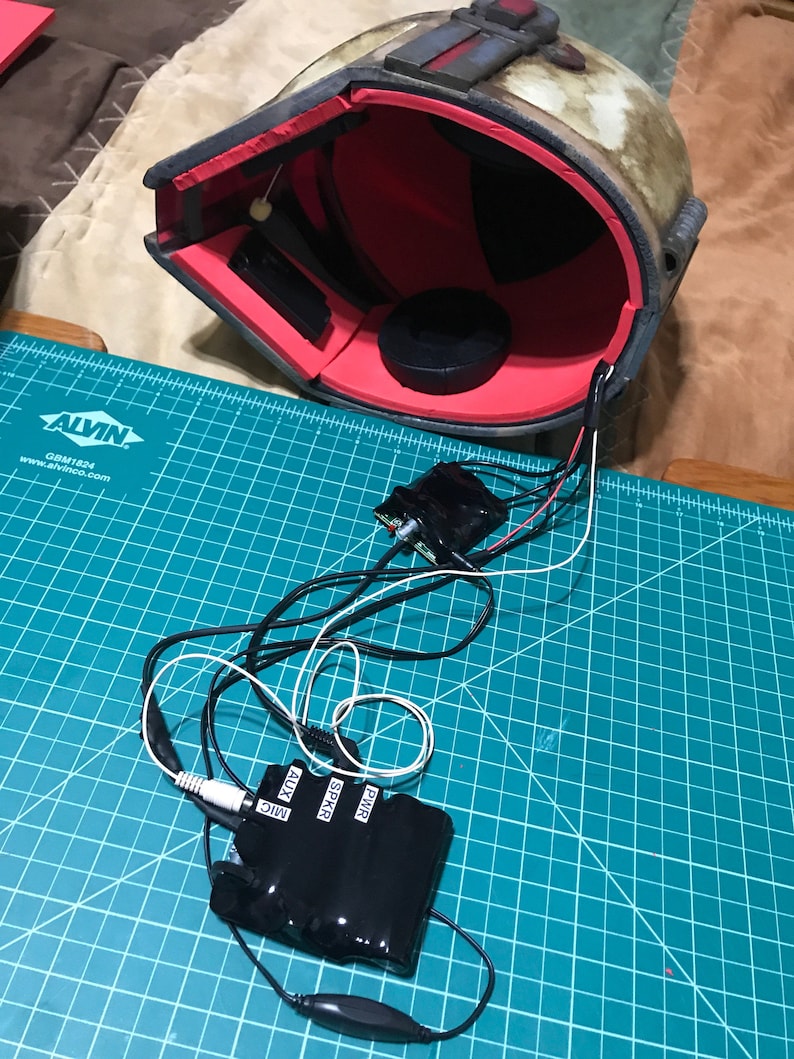 May include: A tan and red helmet interior with black wiring and electronic components. The helmet has a weathered appearance with red padding and a black interior. The wiring is connected to a black box with labels for MIC, AUX, SPKR, and PWR.