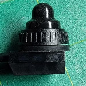 May include: A close-up of a black, plastic component with a rounded top and a ridged, cylindrical base. The component has a rectangular base and is set against a green surface. The object appears to be a small part of a larger mechanism.