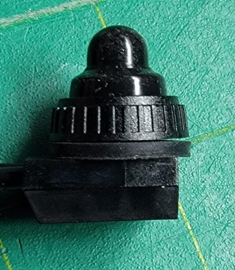 May include: A close-up of a black, plastic component with a rounded top and a ridged, cylindrical base. The component has a rectangular base and is set against a green surface. The object appears to be a small part of a larger mechanism.