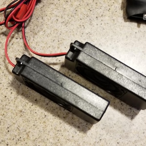 May include: Two black rectangular electronic components with a red and black wire attached. The components are side by side on a speckled surface. The wires are connected to the components.