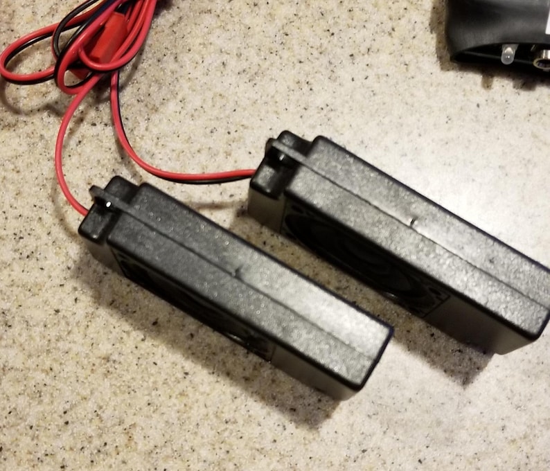 May include: Two black rectangular electronic components with a red and black wire attached. The components are side by side on a speckled surface. The wires are connected to the components.