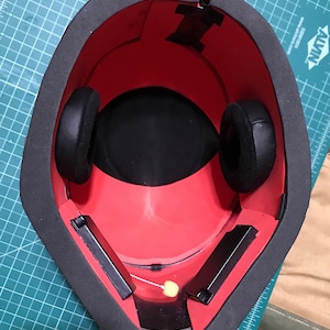 May include: A red and black helmet interior with black ear pads and rectangular black components. The helmet has a thick black border and is set on a green cutting mat. Wires and tape are visible.