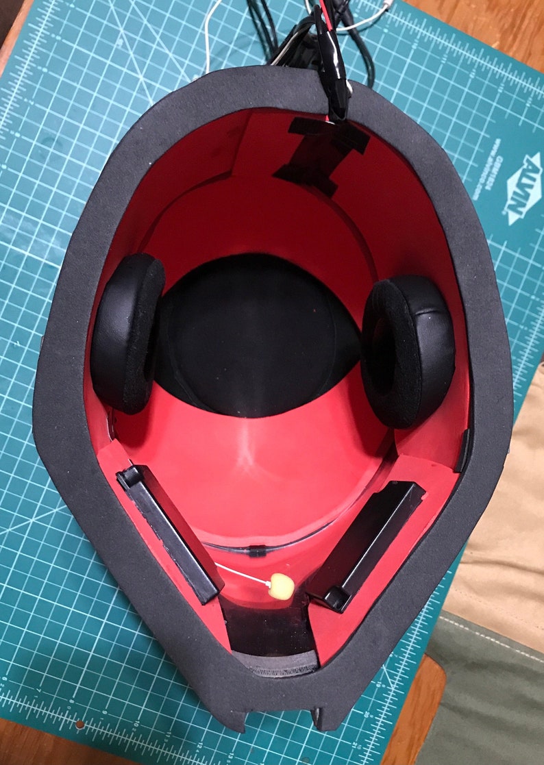 May include: A red and black helmet interior with black ear pads and rectangular black components. The helmet has a thick black border and is set on a green cutting mat. Wires and tape are visible.
