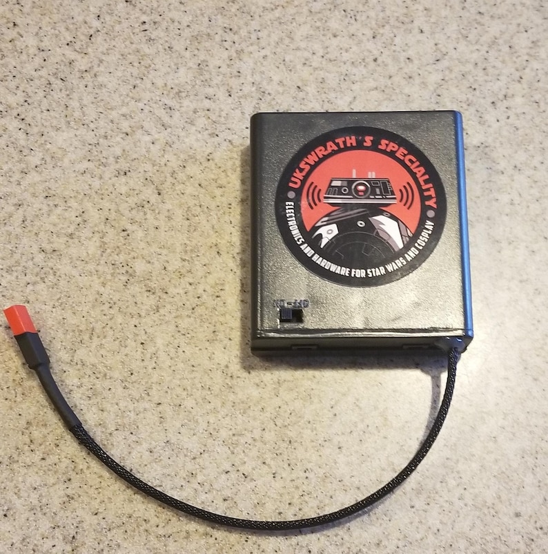 May include: A rectangular, gray electronic device with a black cable and a red connector. The device has a circular sticker with the text "UKSWRATH'S SPECIALITY" and a graphic of a droid. The device also has an on/off switch.