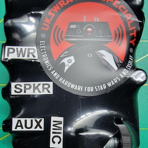 Puede incluir: Caja de plástico negra con una pegatina roja y blanca que dice "Ukswrath's Specialty Electronics and Hardware for Star Wars and Cosplay". La caja tiene etiquetas que dicen "PWR", "SPKR", "AUX" y "MIC".