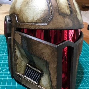 May include: A weathered, brown and tan helmet with a red visor. The helmet has a rectangular vent on the lower left side and a dark frame around the visor. The helmet appears to be made of a durable material.