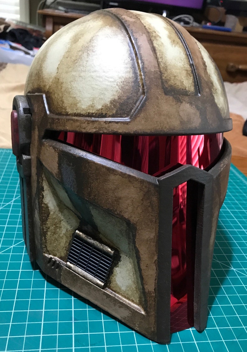 May include: A weathered, brown and tan helmet with a red visor. The helmet has a rectangular vent on the lower left side and a dark frame around the visor. The helmet appears to be made of a durable material.
