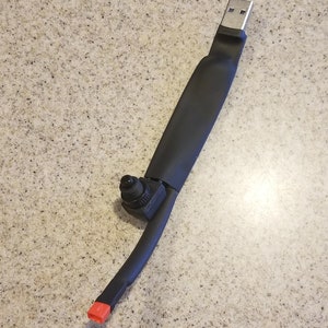 May include: A black USB device with a silver USB connector. The device has a black, rubberized body with a small black component and a red end cap. The device is on a speckled, light brown surface.