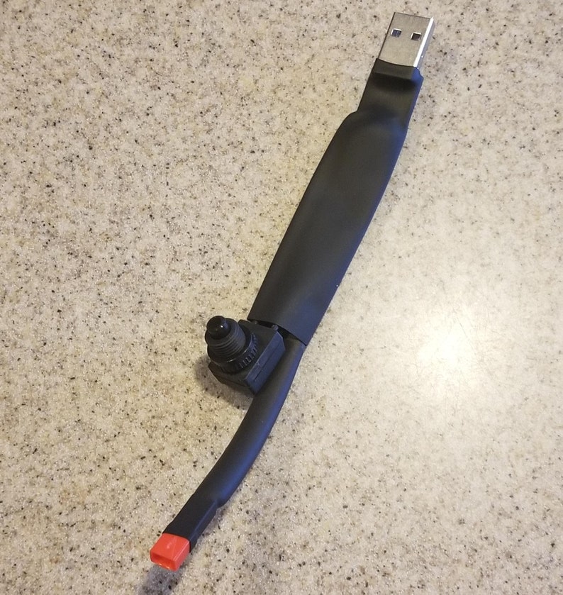 May include: A black USB device with a silver USB connector. The device has a black, rubberized body with a small black component and a red end cap. The device is on a speckled, light brown surface.