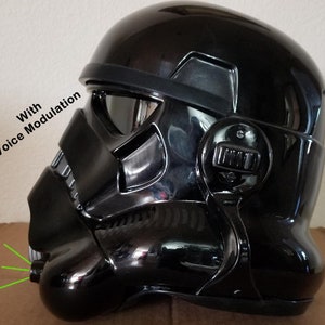 May include: A glossy black helmet with a reflective visor, featuring the text "With Voice Modulation." The helmet has a distinctive design with angular features and a prominent mouthpiece, likely for a costume or collectible.