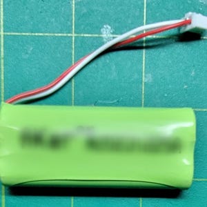 May include: A green cylindrical battery with a white and red wire connected to a white connector. The battery is on a green grid surface.