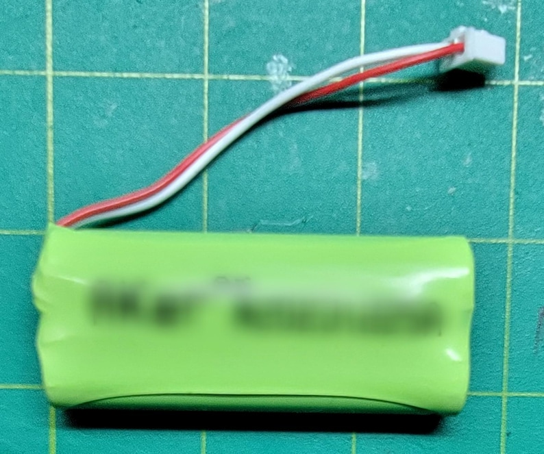 May include: A green cylindrical battery with a white and red wire connected to a white connector. The battery is on a green grid surface.