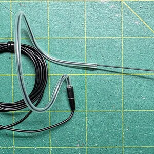 May include: A black headset microphone with a 3.5mm jack, coiled cable, and a clear plastic ear loop. The microphone has a small, black, square foam cover. The background is a green cutting mat.