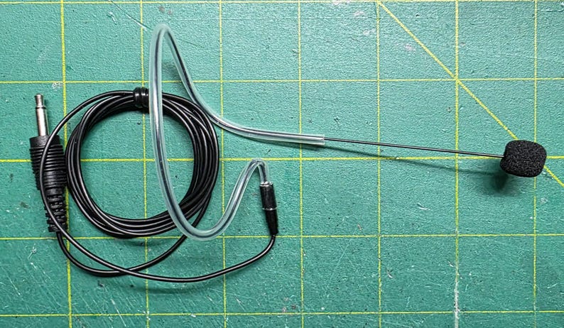May include: A black headset microphone with a 3.5mm jack, coiled cable, and a clear plastic ear loop. The microphone has a small, black, square foam cover. The background is a green cutting mat.