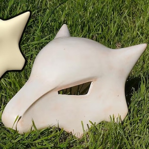 Könnte beinhalten: Zwei weiße Gipsmasken einer mythischen Kreatur mit spitzen Ohren und einem einzigen Augenloch. Die Masken liegen auf einem Bett aus grünem Gras.