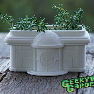 May include: White ceramic planter shaped like a building with a domed roof and a circular window. The planter is filled with green succulents and the text "GEEKYY GARDENS" is visible in the bottom right corner.