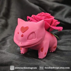 Puede incluir: Un macetero rosa de Pokemon con forma de Eevee con corazones rojos en la cara. Una sola rosa rosa se coloca en el macetero.