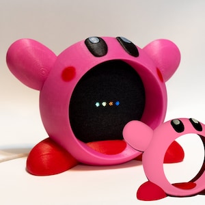 May include: A pink 3D printed  Google Home Mini speaker stand shaped like the video game character Kirby. The stand has a small opening in the front for the speaker's microphone and a row of colorful lights on the speaker.