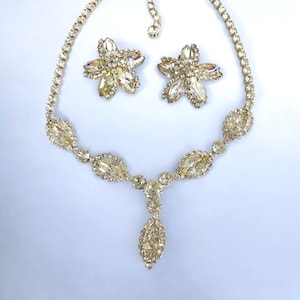 May include: A vintage jewelry set featuring a necklace and matching earrings. The necklace has a series of oval-shaped stones and a central teardrop pendant. The earrings are flower-shaped. All pieces are adorned with clear, sparkling stones.