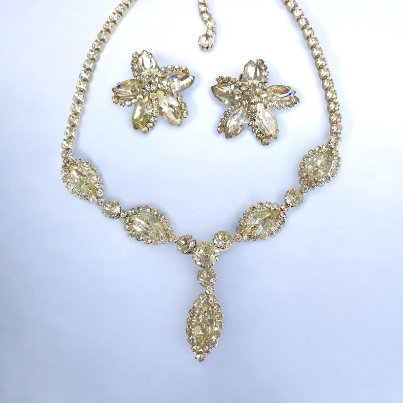 May include: A vintage jewelry set featuring a necklace and matching earrings. The necklace has a series of oval-shaped stones and a central teardrop pendant. The earrings are flower-shaped. All pieces are adorned with clear, sparkling stones.