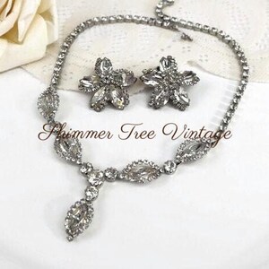 May include: A vintage silver-toned jewelry set featuring a necklace and matching earrings. The necklace has a central pendant and the earrings are flower-shaped. The set is adorned with clear rhinestones. The text "Shimmer Tree Vintage" is visible.