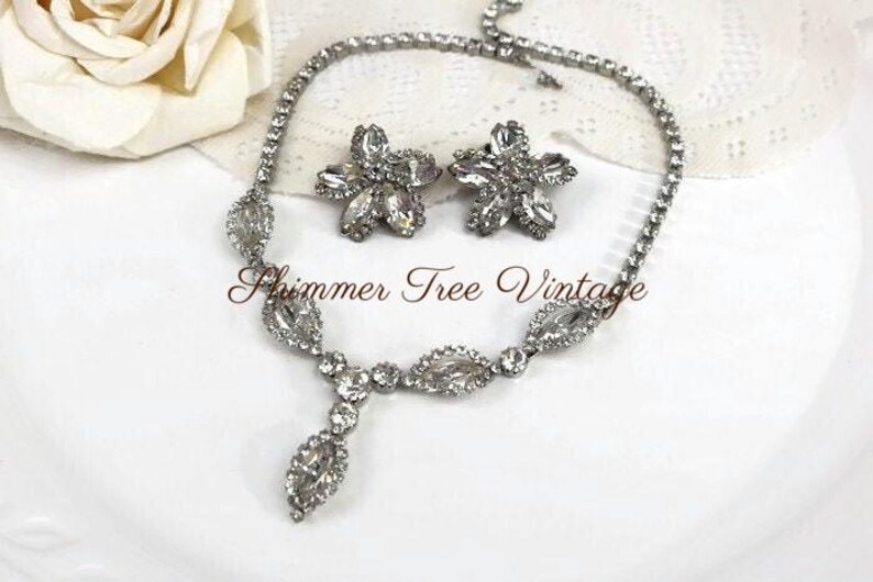 May include: A vintage silver-toned jewelry set featuring a necklace and matching earrings. The necklace has a central pendant and the earrings are flower-shaped. The set is adorned with clear rhinestones. The text "Shimmer Tree Vintage" is visible.