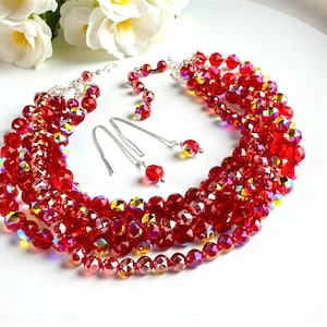 May include: A multi-strand red beaded necklace and matching earrings set. The necklace features several rows of faceted red beads with iridescent highlights. The earrings have a long silver chain with a single red bead at the end. The jewelry is displayed on a white surface.