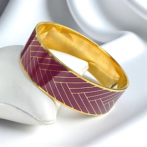 May include: A gold-tone bangle bracelet with a burgundy herringbone pattern. The bracelet has a polished finish and is displayed on a white surface. The design features a repeating geometric pattern, creating a sophisticated look.