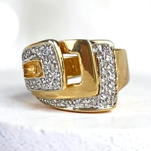 May include: A gold ring featuring a wide, polished band with a buckle-inspired design. The front section is adorned with two rectangular panels encrusted with small, clear, sparkling stones, creating a contrast against the smooth gold surface.