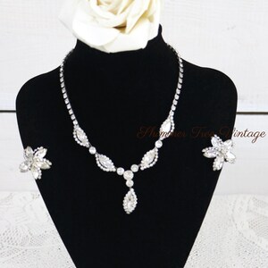 May include: A sparkling silver-toned necklace and matching earrings set on a black display stand. The necklace features a series of clear, faceted stones in a leaf and teardrop design. A large white rose sits atop the stand. The text "Shimmer Tree Vintage" is visible.