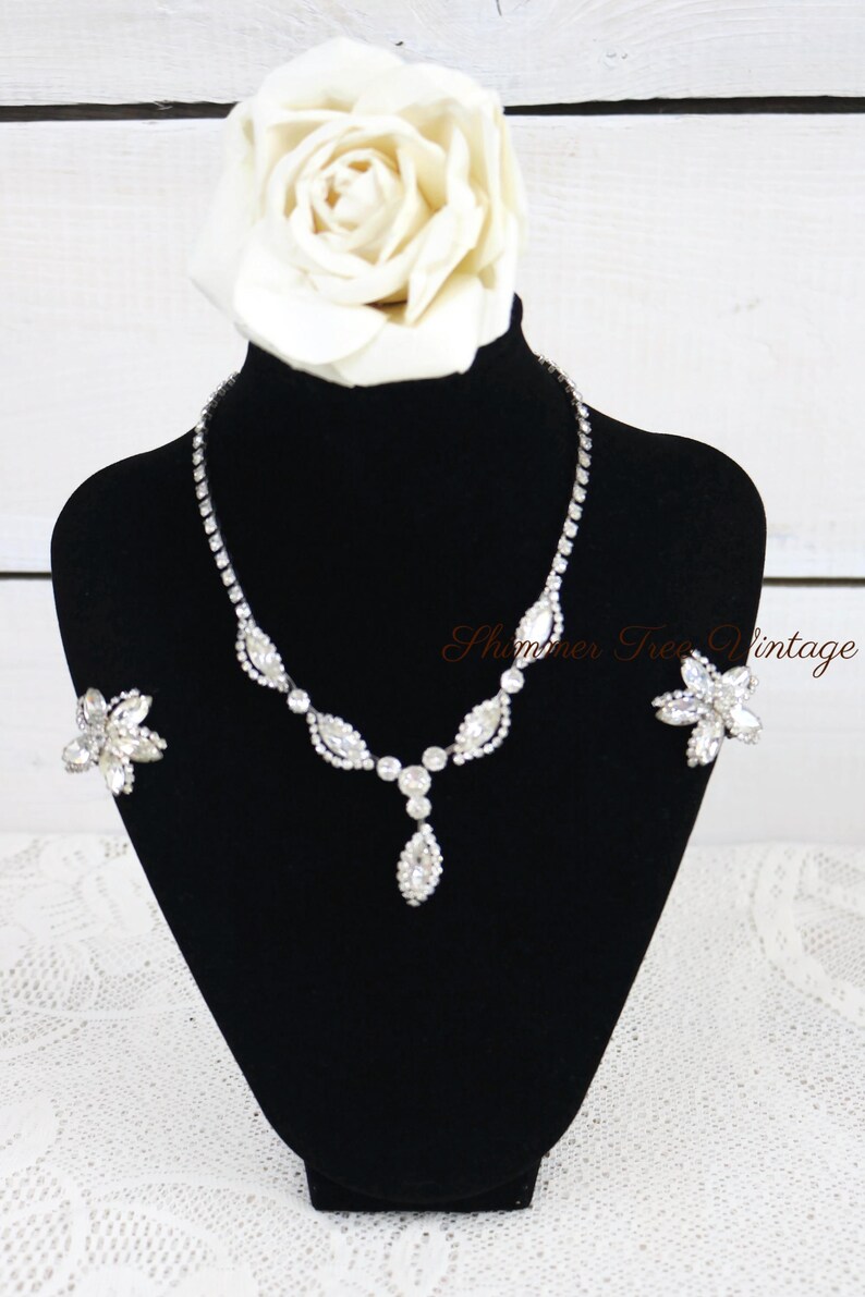 May include: A sparkling silver-toned necklace and matching earrings set on a black display stand. The necklace features a series of clear, faceted stones in a leaf and teardrop design. A large white rose sits atop the stand. The text "Shimmer Tree Vintage" is visible.
