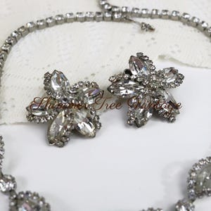 May include: A vintage jewelry set featuring a necklace and matching earrings. The necklace and earrings are silver-toned and adorned with clear, sparkling rhinestones. The earrings are flower-shaped, and the necklace has a delicate design.
