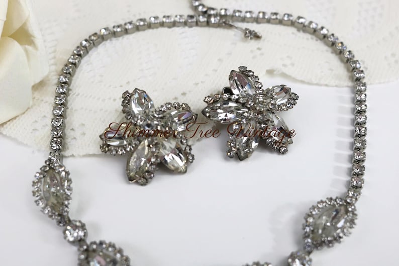 May include: A vintage jewelry set featuring a necklace and matching earrings. The necklace and earrings are silver-toned and adorned with clear, sparkling rhinestones. The earrings are flower-shaped, and the necklace has a delicate design.