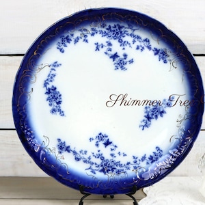 May include: A large, white porcelain plate with a blue and gold floral pattern. The plate has a scalloped edge and is decorated with a blue and gold floral design. The plate is on a white wooden surface.