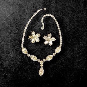 May include: A sparkling jewelry set featuring a necklace and matching earrings. The necklace has a delicate chain with a central pendant. The earrings are flower-shaped. All pieces are adorned with clear, glittering stones, creating a glamorous look.