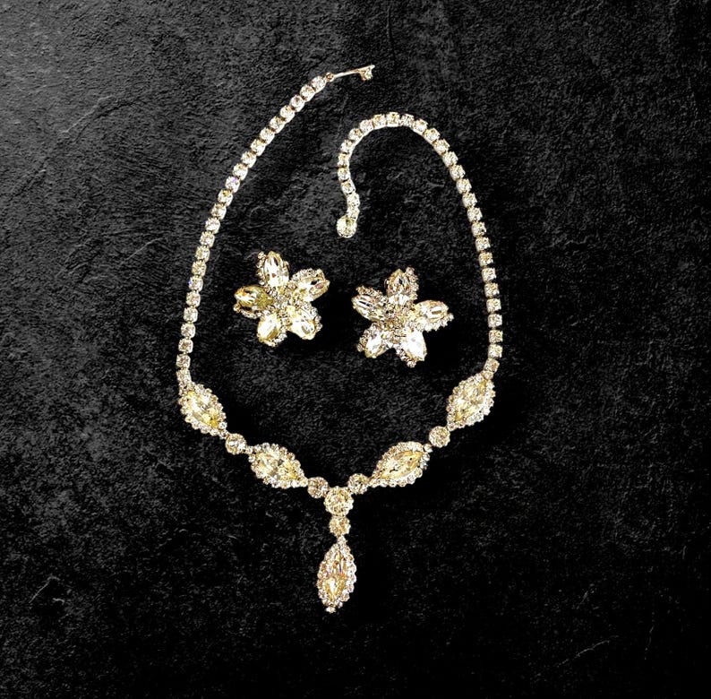 May include: A sparkling jewelry set featuring a necklace and matching earrings. The necklace has a delicate chain with a central pendant. The earrings are flower-shaped. All pieces are adorned with clear, glittering stones, creating a glamorous look.