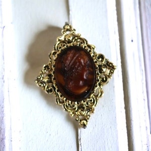 Peut inclure: Une broche dorée à l'or fin avec un design orné présente un camée représentant le visage d'une femme en résine brune.