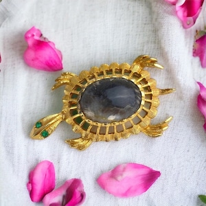 Peut inclure: Une broche en forme de tortue dorée avec un cabochon de pierre grise serti dans un motif de filigrane. La tortue a des accents verts sur la tête et les pattes.
