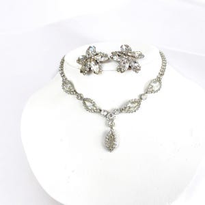 May include: A silver-toned necklace and matching earrings set. The necklace features a series of clear, oval-shaped stones connected by smaller, round stones. The earrings are flower-shaped with multiple clear stones. The jewelry is displayed on a white bust.
