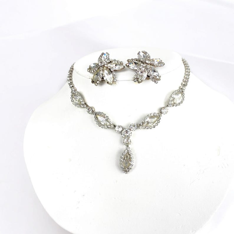 May include: A silver-toned necklace and matching earrings set. The necklace features a series of clear, oval-shaped stones connected by smaller, round stones. The earrings are flower-shaped with multiple clear stones. The jewelry is displayed on a white bust.