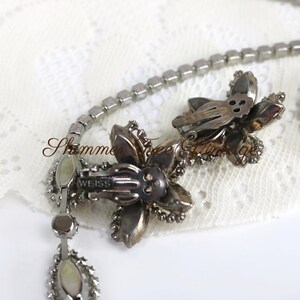 May include: Vintage silver-tone necklace with a floral pendant and a matching pair of flower-shaped clip-on earrings. The necklace features a chain of small, square-cut rhinestones. The earrings and pendant are adorned with sparkling crystals. The brand name "WEISS" is visible.