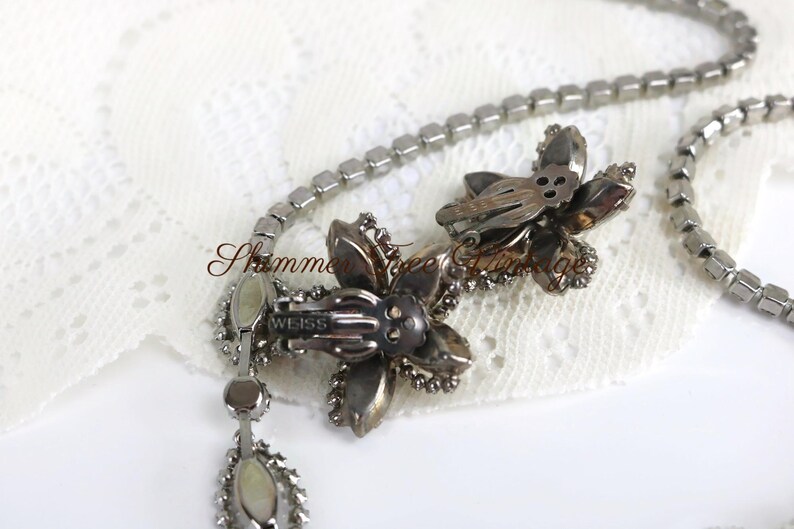 May include: Vintage silver-tone necklace with a floral pendant and a matching pair of flower-shaped clip-on earrings. The necklace features a chain of small, square-cut rhinestones. The earrings and pendant are adorned with sparkling crystals. The brand name "WEISS" is visible.
