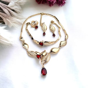 May include: A gold-toned jewelry set featuring a necklace, bracelet, ring, and earrings. The set includes teardrop-shaped red gemstones and small clear stones. The necklace and bracelet have a leaf-like design.