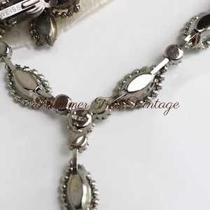 May include: A vintage silver-toned necklace and earring set. The necklace features a series of oval-shaped, clear crystal stones with a teardrop pendant. The earrings are a matching floral design. The brand name "WEISS" is visible on the earring clip.