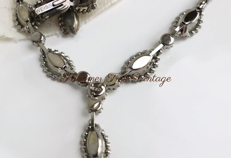 May include: A vintage silver-toned necklace and earring set. The necklace features a series of oval-shaped, clear crystal stones with a teardrop pendant. The earrings are a matching floral design. The brand name "WEISS" is visible on the earring clip.